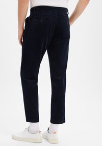Pantalon en velours côtelé bleu marine avec des côtes verticales, coupe slim, deux poches arrière avec boutons, stylé avec des baskets blanches et des chaussettes visibles au niveau des chevilles.