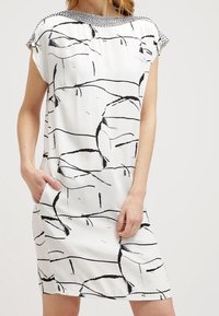 Abito bianco con linee astratte nere, maniche corte e un colletto testurizzato bianco e nero. Presenta tasche laterali e una linea dritta.