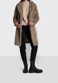 Beige fuskpälsjacka med slag, bärs över en krämfärgad ribbad tröja och svarta jeans. Svarta ankelboots med dragkedjor fulländar outfitten.
