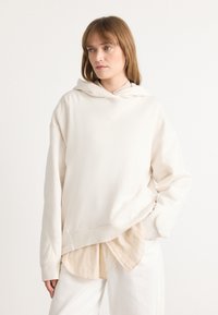 Cream hooded sweatshirt in soft fabric with a relaxed fit, featuring an embroidered logo on the chest and layered over a light shirt.