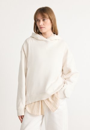 Felpa con cappuccio color crema in tessuto morbido con una vestibilità comoda, caratterizzata da un logo ricamato sul petto e indossata sopra una camicia leggera.