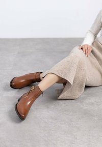 Botas de tobillo de cuero marrón con punta redonda, detalle de hebilla y un tacón bajo y grueso, combinadas con una falda beige texturizada.