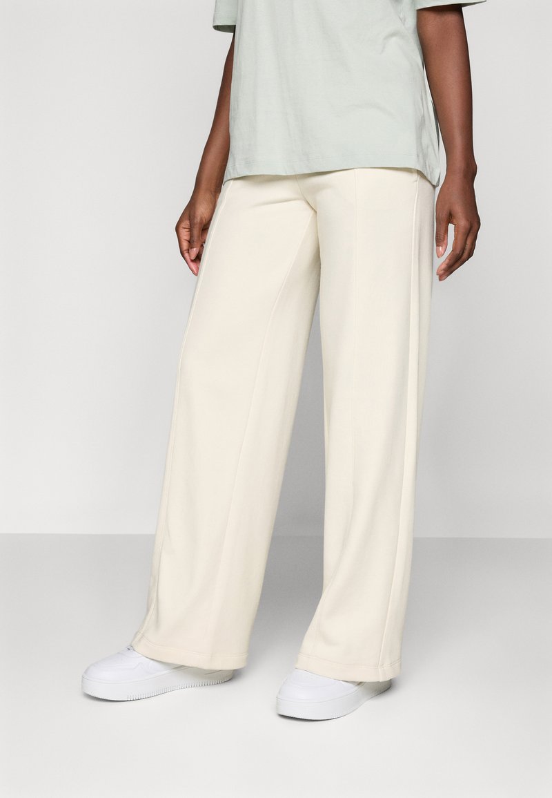 Pantaloni a gamba larga di colore crema, realizzati in tessuto morbido. Abbinati a una cima verde chiaro e leggermente ampia e sneakers bianche. Design minimalista, senza motivi visibili.
