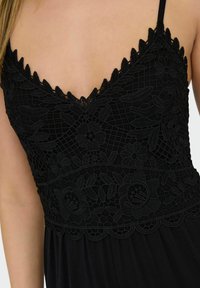 Camisole en dentelle noire avec broderies florales détaillées, décolleté festonné et fines bretelles réglables. Tissu doux et fluide en dessous de la taille.