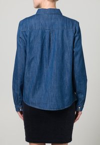 Camicia di denim blu scuro con colletto standard, maniche lunghe e orlo curvo. Presenta pince sul retro e polsini con bottone.