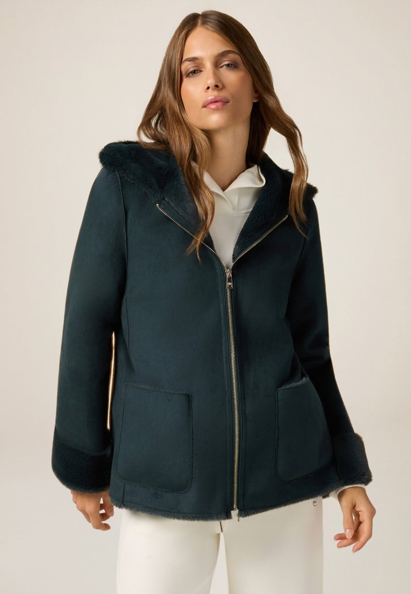 Chaqueta verde oscuro con cremallera y capucha de piel sintética, con puños también de piel sintética, que presenta dos bolsillos frontales y un acabado suave. Diseñada con un corte relajado.