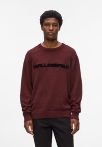 Pullover lavorato a maglia color marrone con collo rotondo, caratterizzato da un logo nero "KARL LAGERFELD" sull'area del petto. Vestibilità ampia con polsini e orlo a costine.