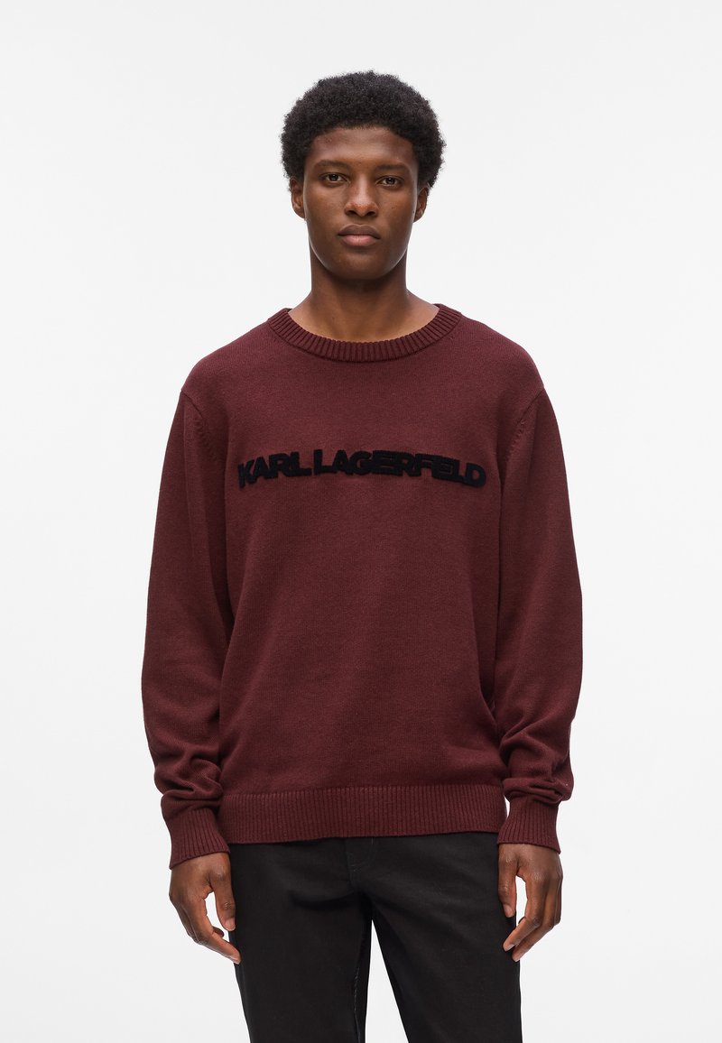 Pullover lavorato a maglia color marrone con collo rotondo, caratterizzato da un logo nero "KARL LAGERFELD" sull'area del petto. Vestibilità ampia con polsini e orlo a costine.