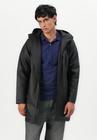 Cappotto impermeabile nero con cappuccio, dotato di chiusura a zip e tessuto testurizzato. Indossato sopra una polo blu navy con pois e jeans grigi.