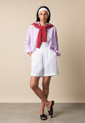 Woman standing with hands in white shorts pockets, wearing lilac shirt, red sweater draped over shoulders, white headband, and black sandals.