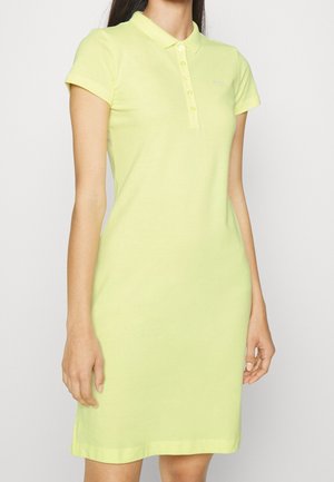 Mujer vestida con un vestido tipo polo de manga corta y color amarillo pálido, con cuello abotonado, de pie frente a un fondo liso y claro.
