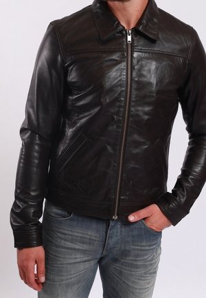 Mand iført en tætsiddende sort læderjakke med lynlås foran og blå jeans, den ene hånd i lommen og den anden afslappet langs siden.