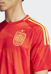 Crvena sportska majica s žutim akcentima, izrađena od teksturiranog materijala, s okruglim izrezom i izvezenim štitastim logotipom na prsima.