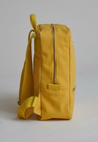 Žlutý batoh vyrobený z odolné tkaniny. Obsahuje svislý zip, boční kapsu a polstrované nastavitelné popruhy pro pohodlí. Obdélníkový tvar.