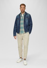 Jeansjacke mit zwei Brusttaschen, grün-blau kariertes Hemd darunter, cremefarbene, schmal geschnittene Hose und weiße Sneakers.