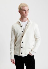 Cardigan color crema con colletto a scialle, bottoni neri, maglia testurizzata e polsini a coste. Indossato sopra una maglietta bianca.