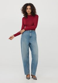 Top rosso a maniche lunghe con collo alto abbinato a jeans chiari a vita alta e scarpe nere con tacco, su uno sfondo bianco semplice.