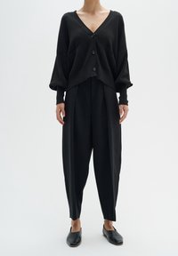 Černý pletený kardigan s výstřihem do V, čtyřmi knoflíky a spadlými rameny. Kombinován s černými širokými kalhotami s vysokým pasem a plochými sandály.