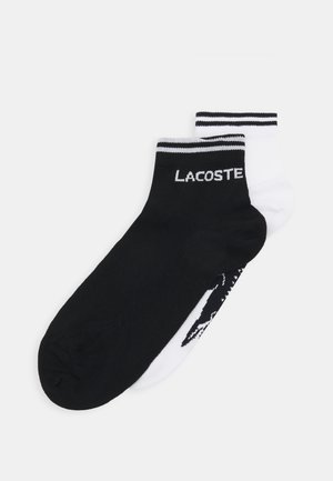 Calcetines tobilleros en blanco y negro, con la parte superior a rayas en contraste y la palabra "LACOSTE" en negrita en el calcetín negro, textura de tela suave.