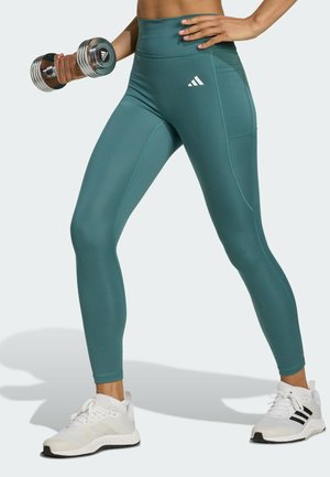 Leggings de sport teal avec une taille haute, dotés d'une poche latérale et d'un logo blanc. Un haltère est tenu dans la main, portés avec des chaussures de sport blanches.