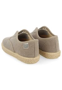 Zapatos slip-on de lona marrón claro con un acabado texturizado, suela de yute trenzado y detalles en metal plateado. La etiqueta trasera presenta el logo de la marca.