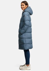 Gevoerde, middellange jas in leisteengrijs met een capuchon. Bevat verticale gestikte secties, zijzakken en een rits sluiting. Gecombineerd met donkere jeans en sneakers.
