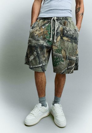 Man wearing camouflage-patterned shorts with drawstring, gray socks, white sneakers, and a gray tank top, standing with hands in pockets.