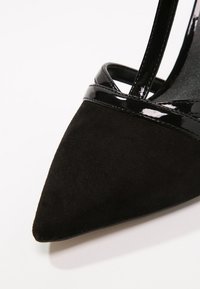 Escarpin noir en daim à bout pointu avec des finitions en cuir verni brillant et une sangle sur fond blanc.