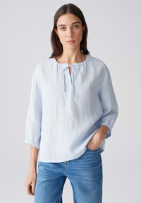 Lichtblauw linnen blouse met een ronde halslijn, strikdetail aan de voorkant en driekwart mouwen, gedragen met denim jeans.