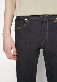 Donkerblauwe denim jeans met oranje stiksels, vijf zakken ontwerp en een knoopsluiting. De stof lijkt duurzaam met een gladde textuur.