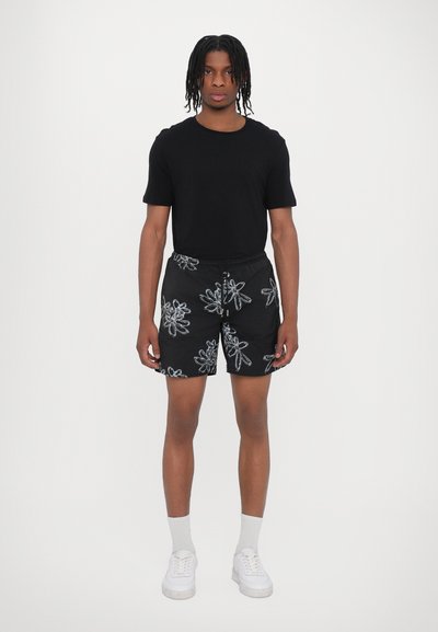 Schwarze Shorts mit einem weißen Blumenmuster, elastischem Bund und Kordelzug. Kombiniert mit einem eng anliegenden schwarzen T-Shirt und weißen Sneakers.