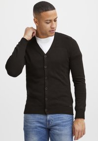 Mørkebrun cardigan, strikket stoff, V-hals design, ribbede skulderdetaljer, knappelukking, brukt over en hvit skjorte, sammen med blåjeans.