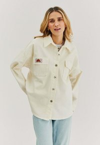 Krémová oversized košile vyrobená z bavlny, obsahující dvě přední kapsy, zapínání na knoflíky a nášivku na levé kapse. Nosič má na sobě světle modré džíny.