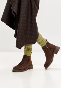 Botas de ante marrón con cordones y cremallera lateral, combinadas con calentadores de piernas acanalados en un tono verde oliva y una falda oscura de rayas diplomáticas.
