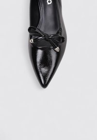 Chaussure noire en vernis avec un bout pointu et une surface texturée, ornée d'un nœud décoratif et d'accents en argent sur le dessus.