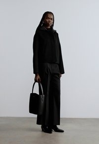 Veste noire ajustée et pantalon noir taille haute, associés à un sac à main noir. Tissu lisse et silhouette moderne, avec des manches longues.
