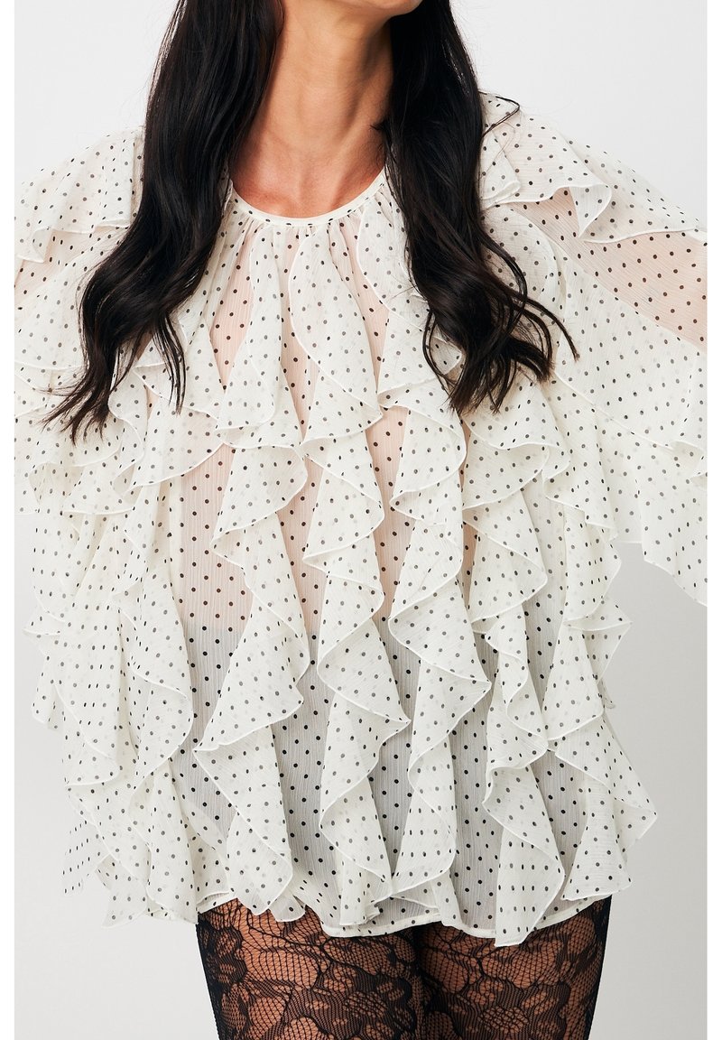Femme portant un chemisier blanc transparent à pois avec plusieurs volants et des collants noirs à motif dentelle, sur fond uni.