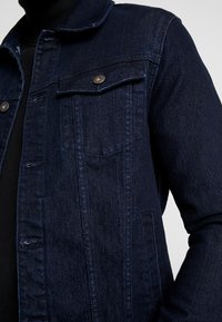 Chaqueta de mezclilla oscura con un cuello clásico, bolsillos frontales y detalles en metal plateado. La textura es suave y tiene un ajuste estructurado.