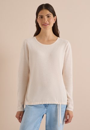 Donna con capelli scuri che indossa un maglione beige chiaro a maniche lunghe con coulisse all'orlo e jeans azzurri, in piedi davanti a uno sfondo semplice.