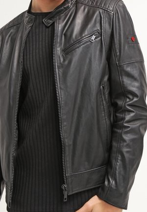 Musta nahkatakki, jossa on vetoketjulliset rintataskut ja sivutaskut, puettuna mustan ribbineulotun paidan päälle. Takki on osittain vetoketjullinen edestä.