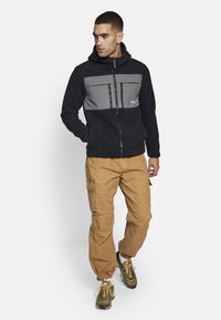 Chaqueta de forro polar negra con panel gris y bolsillos con cremallera, combinada con pantalones cargo color beige y zapatillas multicolor. Modelo posado frente a un fondo neutro.