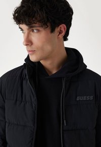 Sort quiltet pufferjakke med lynlås, med høj krave og hætte, struktureret stof og et trykt "GUESS" logo på brystet.