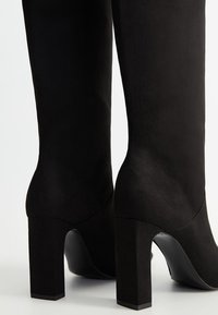 Bottes noires montantes en suède avec une texture lisse. Dotées d'un talon épais et carré et d'un design de bout arrondi.