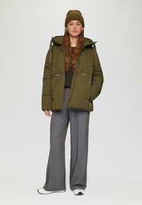 Groene pufferjack met rits en trekkoord, met twee voorkruimelpockets. Gecombineerd met grijze wijdvallende broek en een bruin gebreid mutsje.