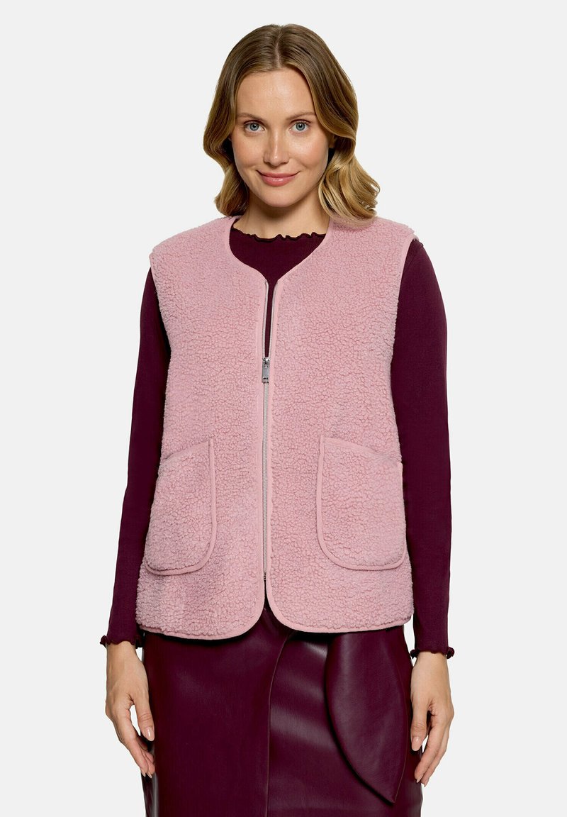 Femme portant un gilet polaire rose clair zippé sur un haut à manches longues et une jupe bordeaux, debout face à un fond uni.