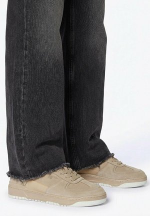 Beige suède sneakers met veters gedragen bij brede zwarte jeans met rafelige zoom, tegen een effen witte achtergrond.