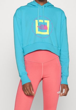 Person iført en cropped turkis hoodie med en gul og pink The North Face-logo og koralrosa leggings med et lille logo på låret.
