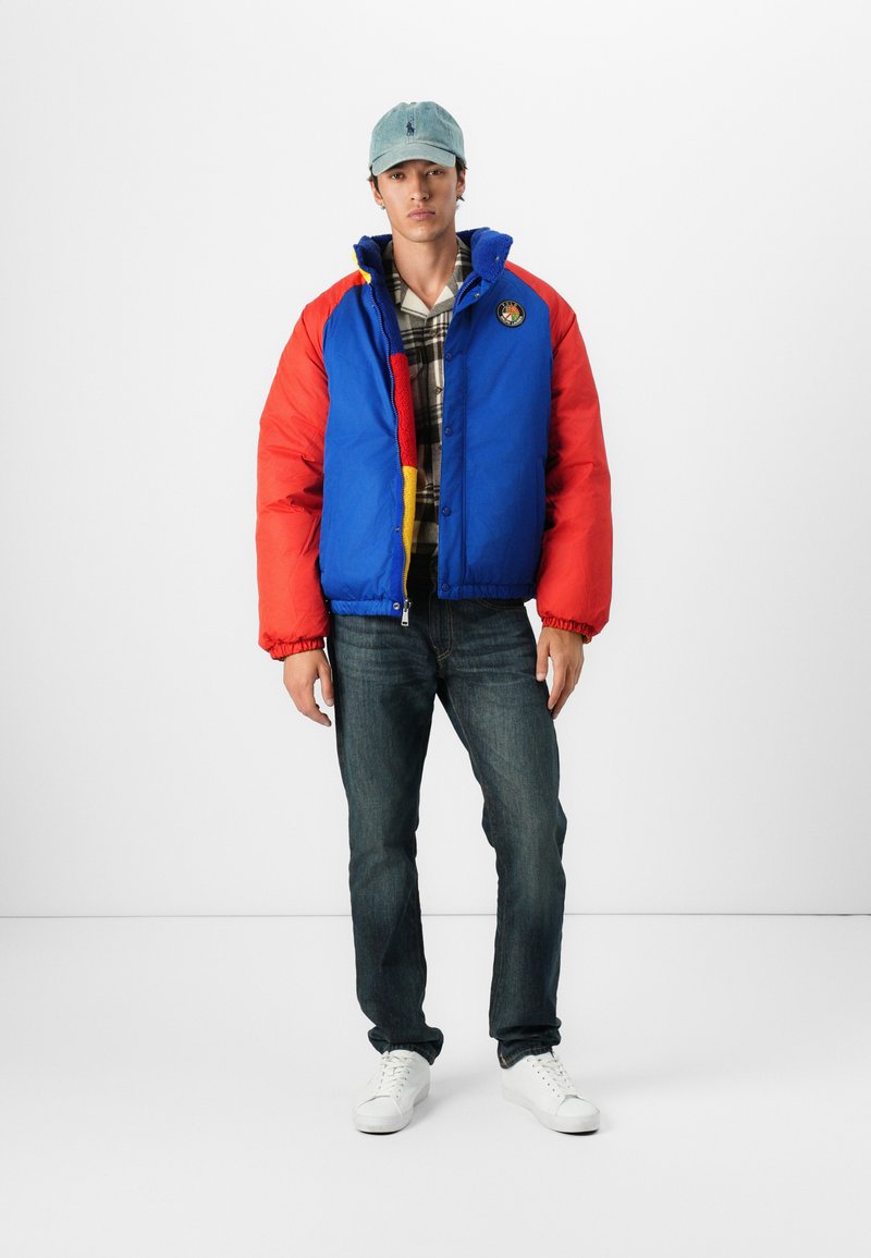 Kolorowa kurtka puchowa z niebieskimi i czerwonymi panelami, okrągłym logo na klatce piersiowej, zestawiona z koszulą w kratę, ciemnymi dżinsami i białymi trampkami.