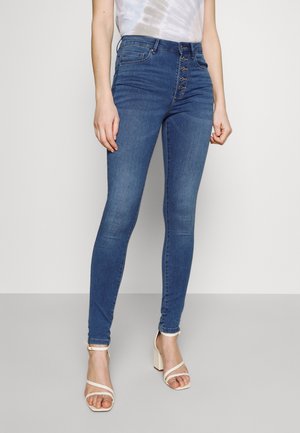 Slim-fit blauwe jeans met hoge taille en knoopsluiting, gedragen met witte bandjes sandalen met blokhak en een licht tie-dye top.