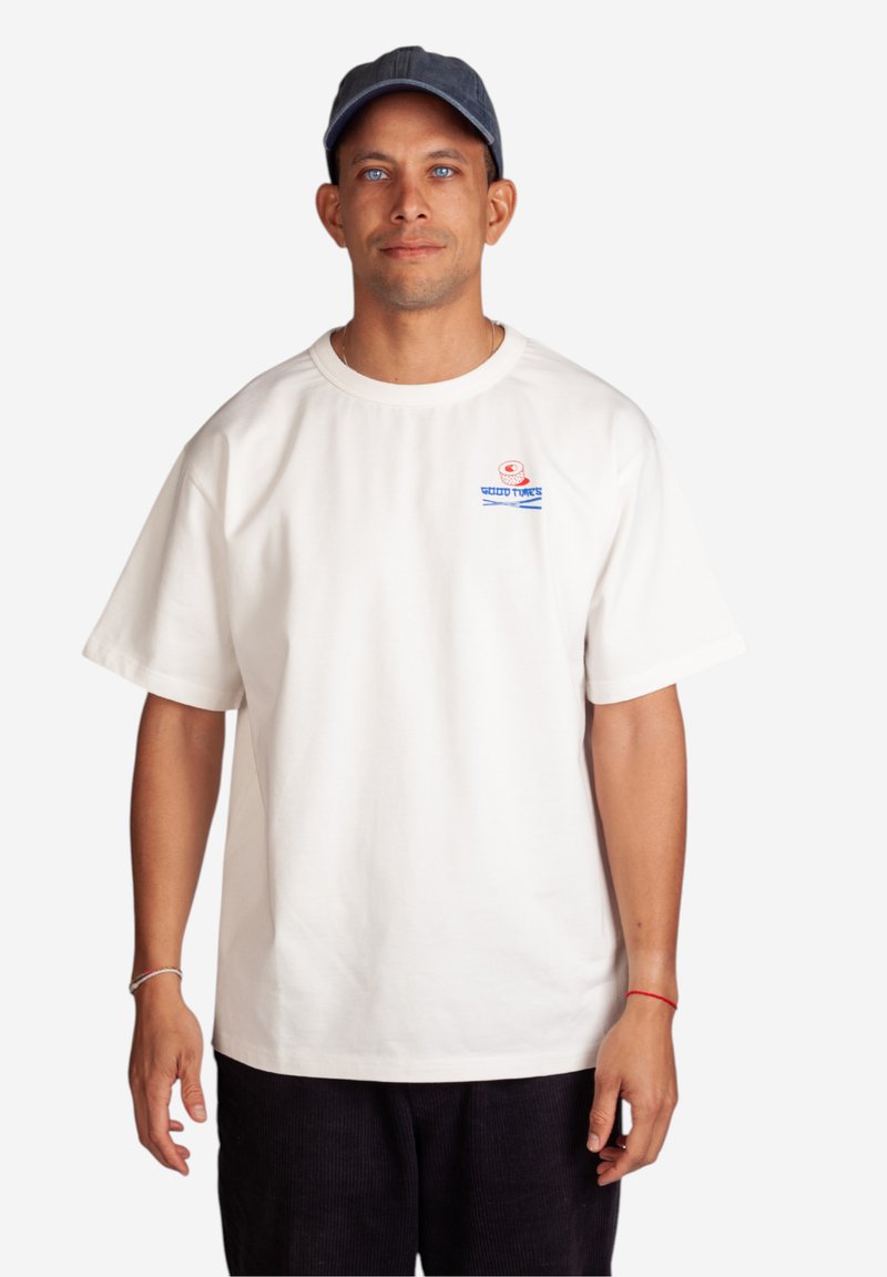 Witte katoenen t-shirt met een ronde hals, korte mouwen en een blue grafisch ontwerp op de borst. Gekocht met donkere corduroy broeken en een marineblauwe pet.